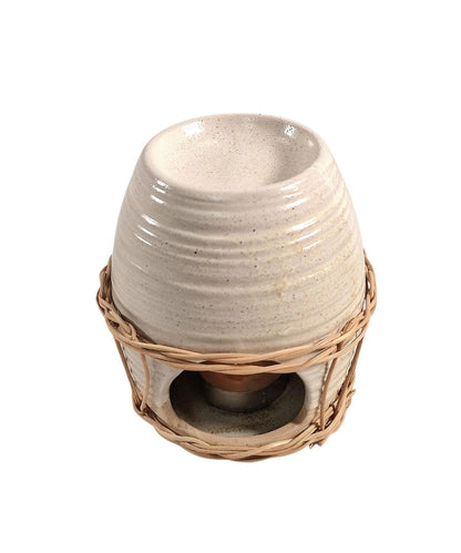 Ceramic oil burner with natural rattan detail, featuring a lit tealight inside, styled on a wooden surface with a floral arrangement in the background