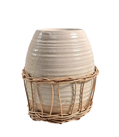 Ceramic oil burner with natural rattan detail, featuring a lit tealight inside, styled on a wooden surface with a floral arrangement in the background