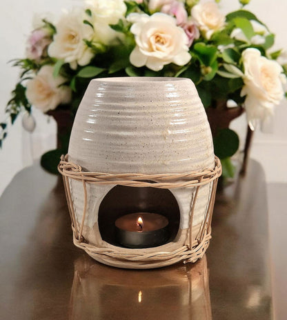 Ceramic oil burner with natural rattan detail, featuring a lit tealight inside, styled on a wooden surface with a floral arrangement in the background